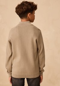 Maglione beige in maglia con collo alto e polsini a coste. Tessuto liscio, vestibilità comoda e punti sottili sulle spalle. Pantaloni scuri visibili sotto.