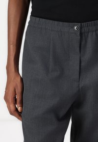 Grijze stoffen broek met een gestructureerde weving, elastische tailleband en een enkele knoopsluiting. Hand zichtbaar aan de zijkant.
