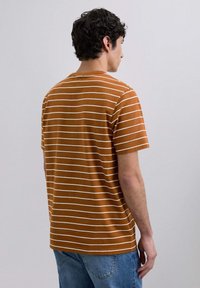 Camiseta de rayas marrón con líneas horizontales blancas, hecha de un tejido suave. Mangas cortas y escote redondo. Se lleva con vaqueros azules.
