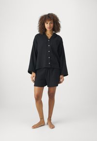 Schwarzes Baumwoll-Loungewear-Set mit einer locker sitzenden, langen, knöpfbaren Bluse mit Tasche und passenden Shorts, beide mit einer feinen Textur.