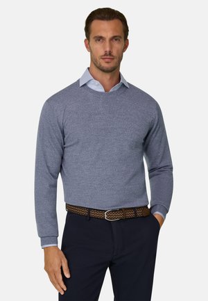 Homme portant un pull texturé bleu clair par-dessus une chemise à col, un pantalon foncé et une ceinture tressée marron, debout avec une main dans la poche.