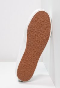 Baskets blanches avec une semelle extérieure en caoutchouc texturée. Vue de côté montrant une tige lisse et un bord renforcé. Motif d'adhérence détaillé sur la semelle.