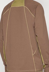 Long-sleeve shirt in brown fabric with yellow stitching. Features a textured upper panel and seam accents along the sleeves and sides.