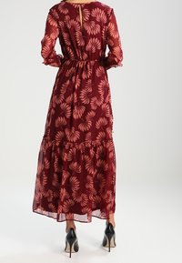 Robe florale bordeaux avec manches longues en voile, taille froncée, jupe à volants et détail clé dans le dos. Portée avec des chaussures à talons noires.