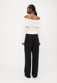 Off-Shoulder gerippter weißer Top, kombiniert mit schwarzen weiter geschnittenen Hosen. Das Top hat lange Ärmel und einen lockeren Sitz. Glatte Stofftextur.