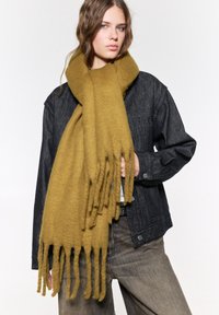 Écharpe en laine jaune avec des franges, portée lâchement autour du cou, associée à une veste en denim foncé et un pantalon gris.