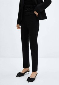 Pantalon noir ajusté en tissu lisse, coupe slim avec une taille mi-haute, associé à des chaussures à l'arrière découvert en noir avec un détail en nœud.