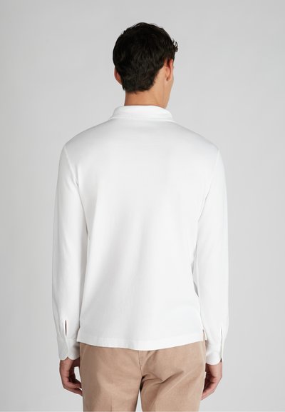 Uomo che indossa una polo bianca a maniche lunghe e pantaloni beige, in piedi con le spalle rivolte alla camera su uno sfondo grigio uniforme.
