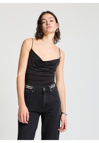 Schwarzer Bodysuit mit engem Kragen, schmalen Trägern und dekorativem Schmuck am linken Schulterbereich, kombiniert mit hoch taillierten schwarzen Jeans mit Verzierungen.