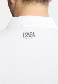Maglietta polo bianca con tessuto testurizzato. Presenta un colletto e un logo grigio: "KARL LAGERFELD BEACHWEAR" sul retro.