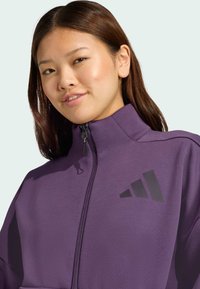 Fialová zipsová bunda vyrobená z mäkkej látky s vysokým golierom. Obsahuje čierne logo na hrudi. Hladká textúra, priliehavý strih.