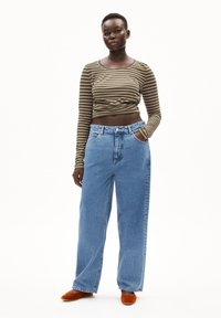 Gestreiftes Langarmoberteil in Olivgrün und Braun, kombiniert mit blauen weitem Jeans. Das Model trägt orangefarbene Slipper. Einfacher, lässiger Look.