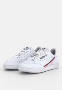 Pair of white Adidas sneakers with black and red logo on side, white laces, and perforated toe caps on a plain background.