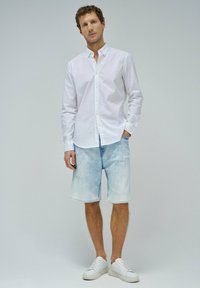 Camisa blanca de botones, pantalones cortos de denim desgastados de color azul claro y zapatillas blancas. La camisa tiene un cuello y botones en el frente.