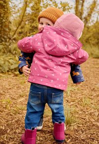 Dos niños se abrazan al aire libre. Uno lleva una chaqueta de lluvia rosa con flores, mangas azul con patrones y botas rosas. El otro lleva un gorro rosa.