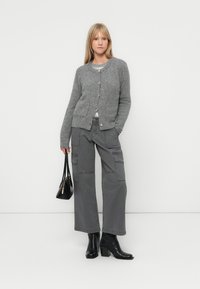 Grå strikket cardigan med knapper, kombinert med grå cargo-bukser med lommer. Modellen bærer en liten svart håndveske og har på seg svarte hæler.