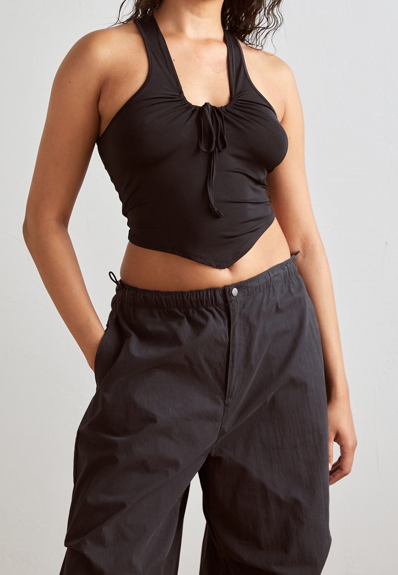 Femme portant un crop top noir sans manches avec un détail noué et un pantalon noir ample, debout avec une main dans la poche sur un fond uni.
