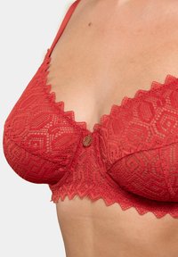 Soutien-gorge en dentelle rouge avec un bord festonné, présentant des motifs géométriques et un accent métallique décoratif au centre. Bretelles ajustables.
