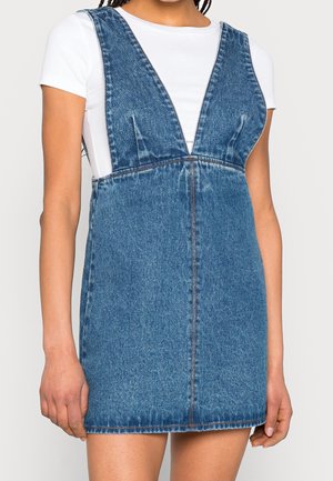 Femme portant une mini-robe en jean bleu sans manches avec un décolleté plongeant en V par-dessus un top blanc ajusté à manches courtes.
