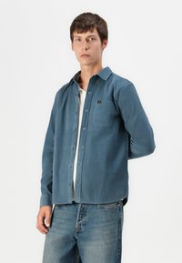 Blue button-up shirt made of a soft fabric, featuring a chest pocket and a relaxed fit. Paired with light blue denim pants.
