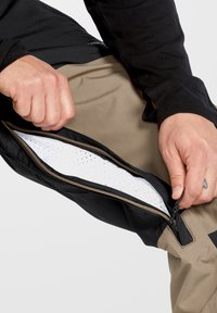Zwarte en beige broek met een ritszak; binnenwerk heeft een witte mesh voering; de hand van een persoon past de rits aan.