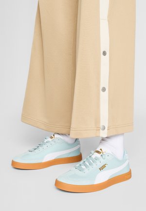 Lichtblauwe sneakers van suede en leer, met witte accenten en oranje rubberen zolen; gecombineerd met beige wijde broek met drukknoopsluiting.