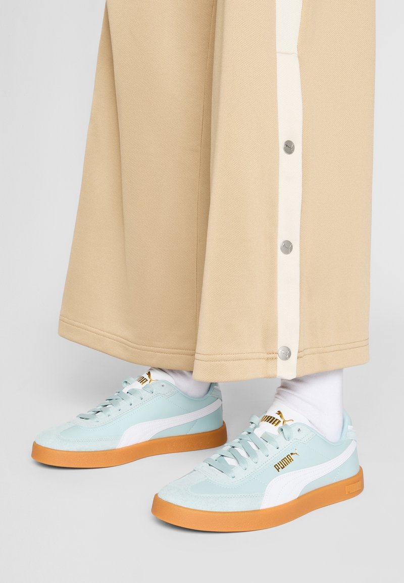 Sneaker azzurri chiari con materiali in suede e pelle, dettagli bianchi e suole in gomma arancione; abbinati a pantaloni beige a gamba larga con bottoni.