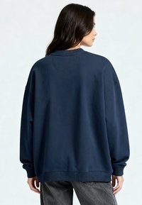 Femme aux longs cheveux bruns, portant un sweat-shirt bleu marine oversize et un jean noir délavé, debout, tournée vers la droite, de dos.