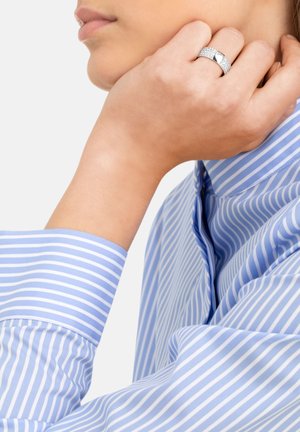 Anello d'argento con pietre trasparenti, caratterizzato da un design a banda liscia. Indossato su una mano che poggia su una camicia a righe blu e bianche.