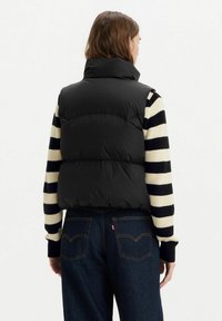 Schwarweste mit hohem Kragen, kombiniert mit einem schwarz-creme gestreiften Pullover, getragen über dunklen Jeans.