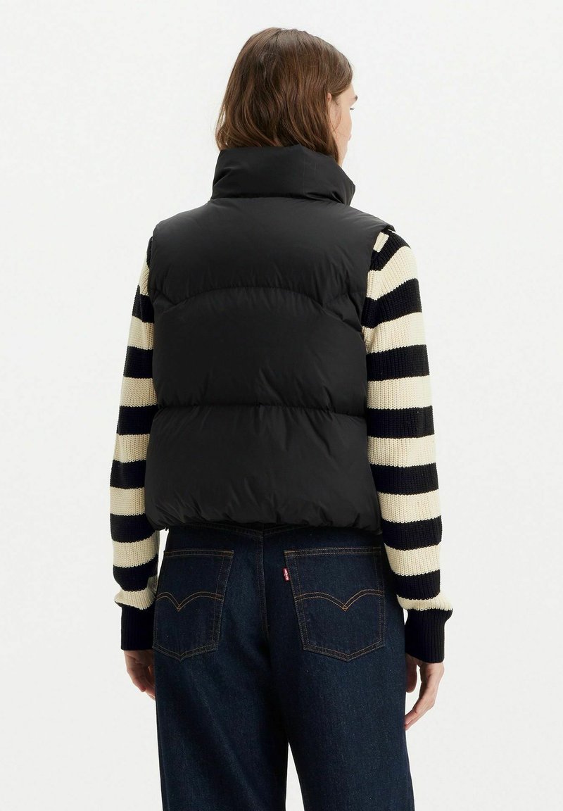 Gilet imbottito nero con colletto alto, abbinato a un maglione a righe nere e crema, indossato su jeans in denim scuro.