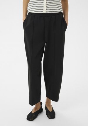 Persoon die zwarte cropped losse broeken draagt, zwarte gebreide slip-on schoenen en een mouwloos wit topje met zwarte strepen.