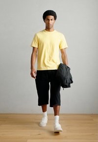 Κίτρινο κοντομάνικο μπλουζάκι, μαύρο σορτς μέχρι το γόνατο, λευκά αθλητικά παπούτσια και σκούρο μπουφάν κρατημένο στο αριστερό χέρι, στέκεται πάνω σε ξύλινο δάπεδο.