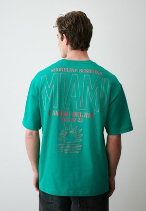 Hátrafelé néző férfi zöld pólóban, piros felirattal: „Shoreline Sessions MIAMI Camino Del Rey Aug 20-23“, alatta nap grafikával.