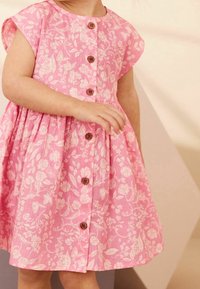 Růžové šaty s květinovým potiskem a krátkými rukávy, s předním zapínáním na knoflíky z dřeva a širokou sukní. Jsou viditelné detaily texturované tkaniny.