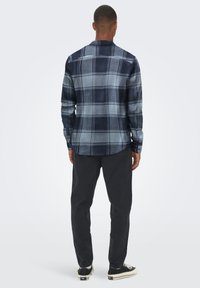 Hombre vestido con una camisa de cuadros azul marino y celeste de manga larga, pantalones negros y zapatillas en blanco y negro, de pie mirando hacia atrás.