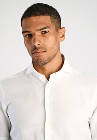 Jonge man met kort, krullend haar en lichte gezichtsbeharing, gekleed in een wit overhemd met knopen, tegen een lichte effen achtergrond.