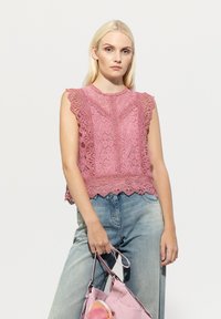 Rosa Spitzenoberteil mit Rüschensleeves, aufwendigen Mustern und einem gewellten Saum, kombiniert mit hellblauen Jeans und einer rosa Tasche.