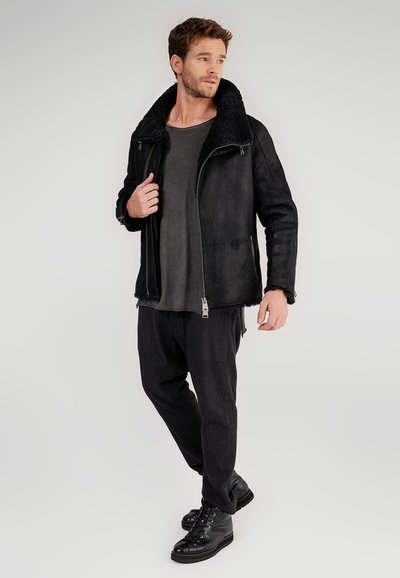 Uomo che indossa una giacca di shearling nera, una maglietta grigio scuro, pantaloni neri e stivali neri con lacci, in piedi contro uno sfondo semplice.