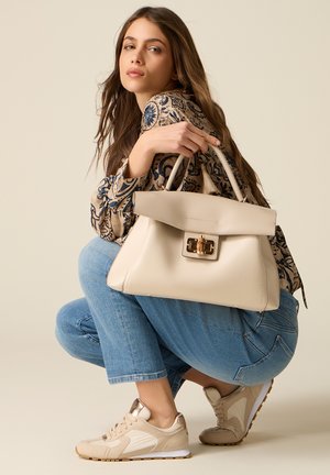 Junge Frau hockt, trägt eine gemusterte Bluse, blaue Jeans und beige Sneakers, hält eine große beige Handtasche mit goldenem Verschluss.