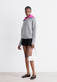 Grijze gebreide trui met een roze kraag, gecombineerd met een zwarte rok. Houdt een kleine witte handtas vast en draagt zwarte ballerina's met een sluiting.