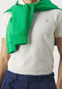 Man in witte polo met geborduurd logo, groene sweatshirt over de schouders gedrapeerd, en navyblauwe geplooide broek.