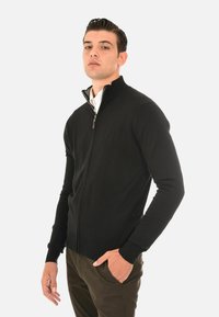 Maglione nero con zip, colletto alto, polsini a costine e design aderente. Indossato su una camicia con colletto, mostrato su un modello.