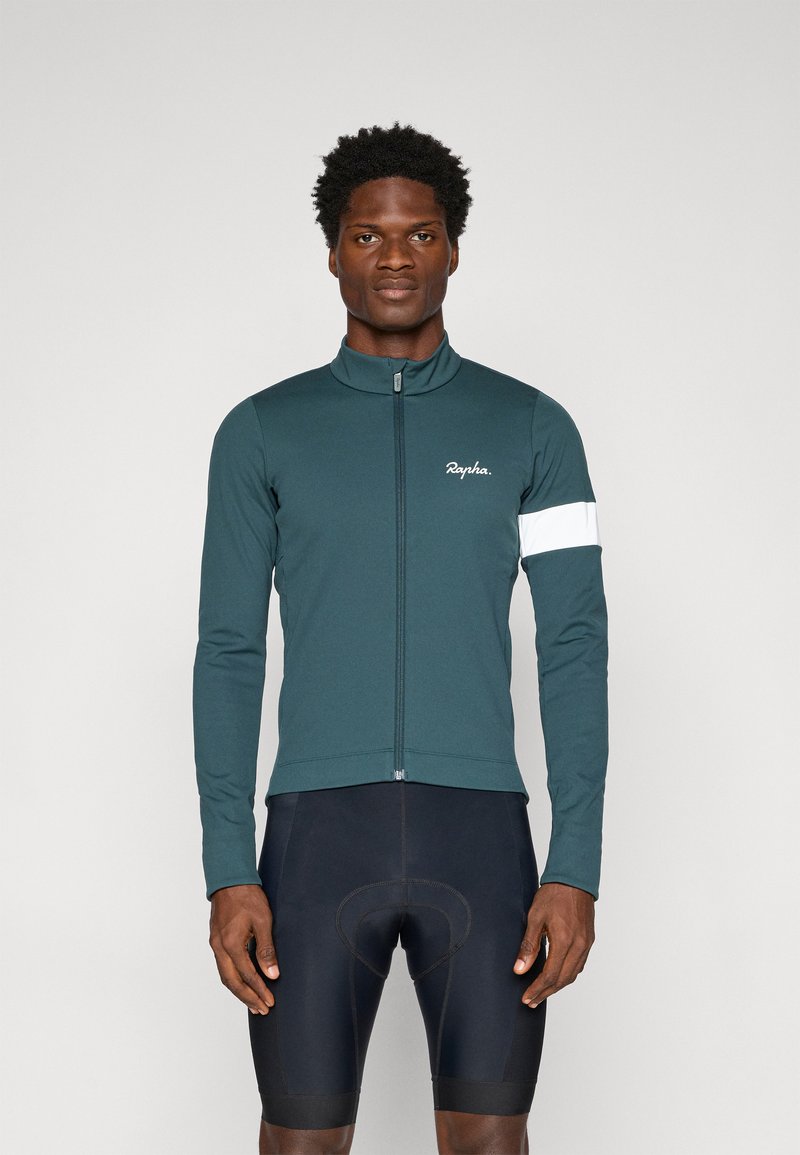 Chaqueta de ciclismo de manga larga en tela de color teal oscuro, con cremallera frontal, cuello alto y una banda de acento blanca en el brazo izquierdo. Pantalones cortos negros.