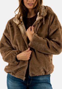 Chaqueta de pelaje sintético marrón con una textura suave, cremallera frontal y capucha. Forrada con una tela de color claro. Combinada con una blusa oscura.