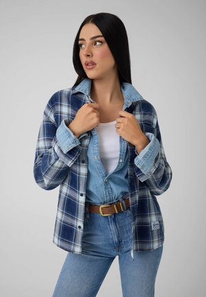Femme aux longs cheveux noirs portant une chemise à carreaux bleue par-dessus une veste en jean, un haut blanc et un jean bleu, regardant de côté sur un fond uni.