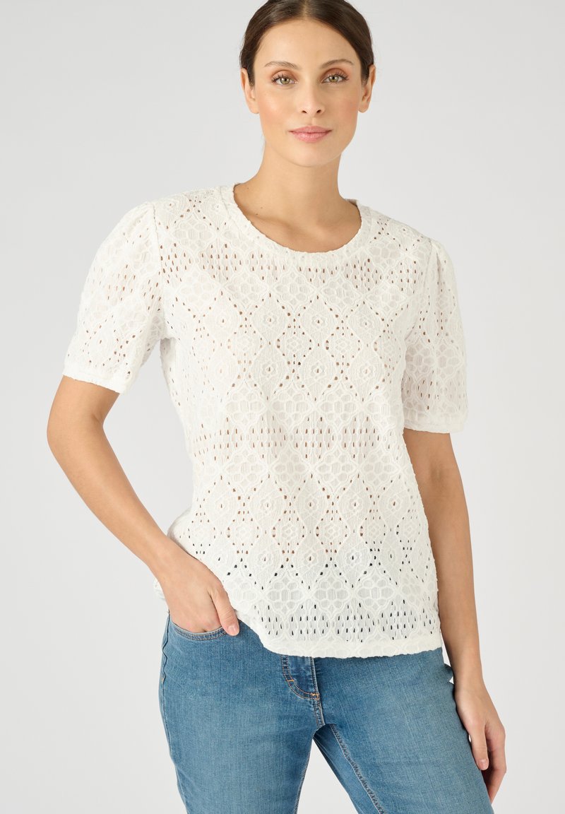 Witte katoenen blouse met korte pofmouwen, voorzien van intricates diamant-gatpatronen en een ronde halslijn. Gecombineerd met blauwe jeans.