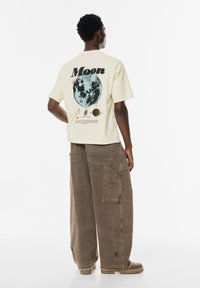Lichtgele t-shirt met een afbeelding van de maan en planeten op de rug, gecombineerd met losse bruine cargobroek met zakken.