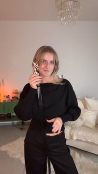 Μαύρο τοπ με μακριά μανίκια και μαύρο παντελόνι, κρατώντας μια διακοσμητική αγκράφα ζώνης. Απαλές, φωτεινές υφές φόντου με ζεστό φωτισμό.