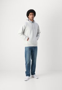 Hanorac cu glugă de culoare gri deschis, cu buzunar frontal; purtat cu blugi albastre și adidași albi. Modelul stă cu spatele la un fundal neutru.